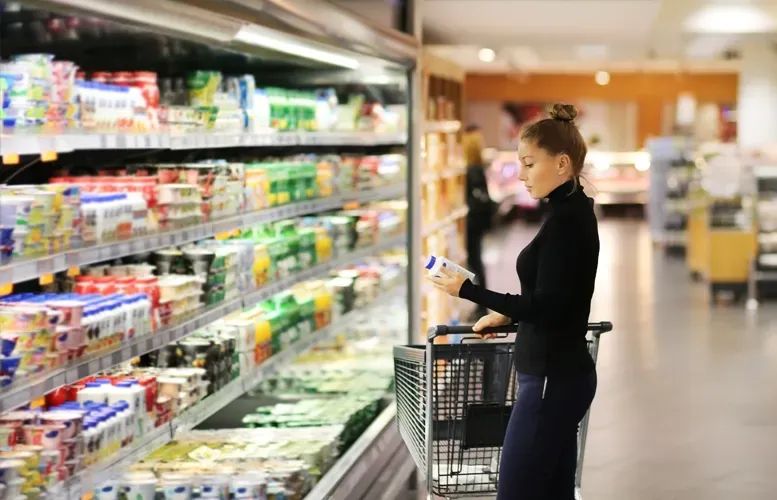 Woman shopping, looking at yogurt in a refrigerated grocery display, with a shopping cart in a store aisle.