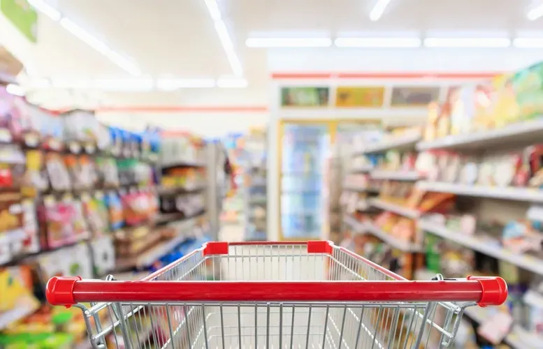 Shopping cart in a brightly lit grocery store aisle, looking at shelves of products.