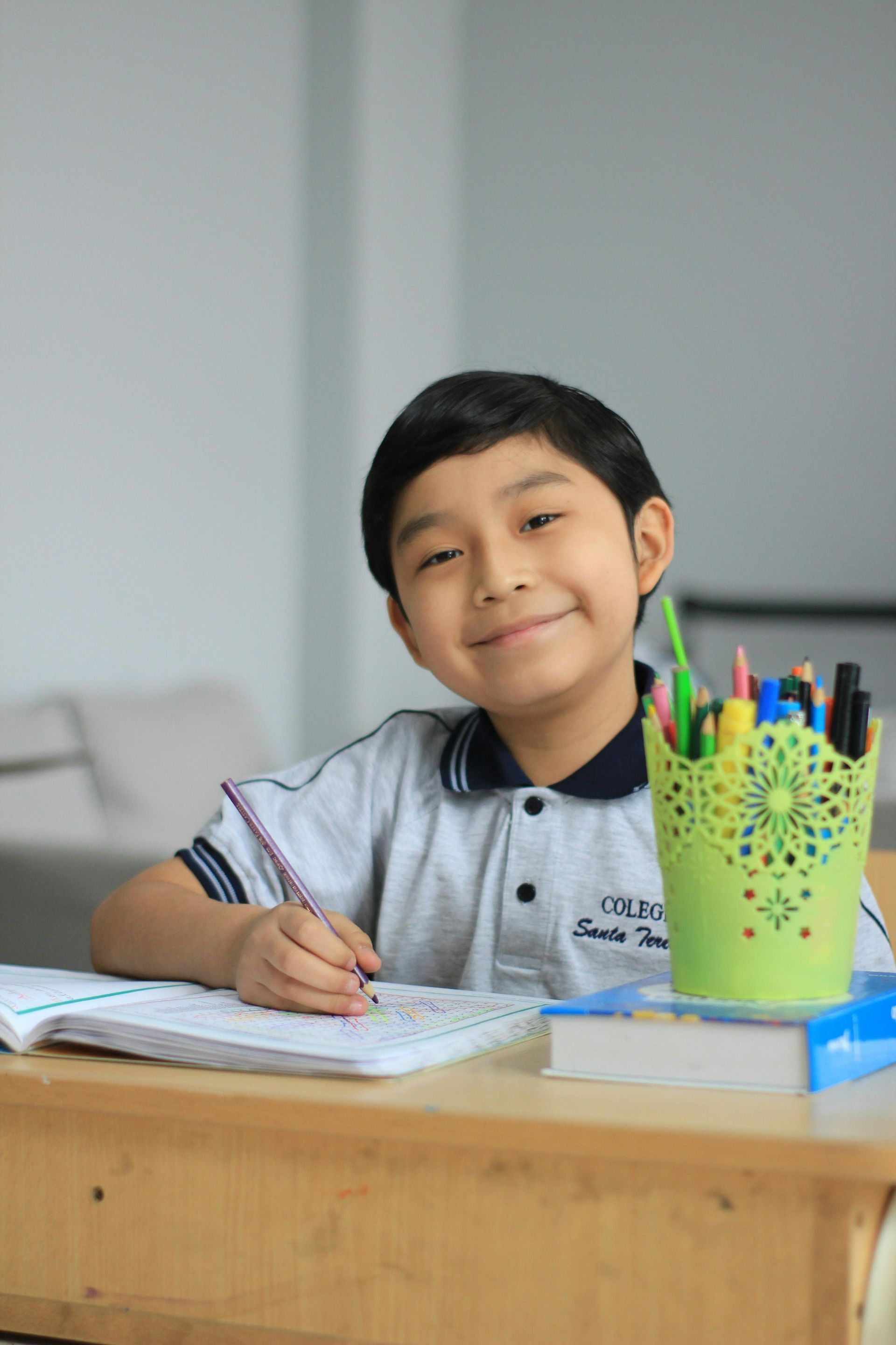 Boy smiling, writing in notebook at desk, pencil holder with colorful pens and pencils.
