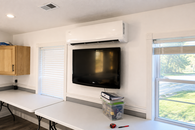Bright room with wall-mounted TV, air conditioner, folding tables, windows, and a wooden cabinet.