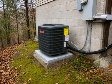 Outdoor air conditioner unit beside a house on a concrete pad, with fallen leaves and woods nearby