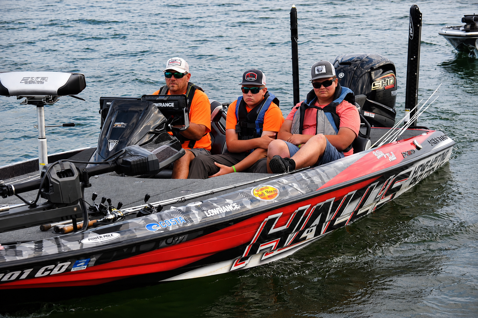 Four anglers in life vests ride a red-and-black HAWG bass boat on calm water.