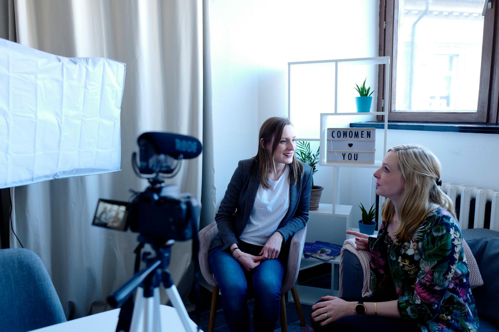 Two women are sitting in front of a camera in a living room.