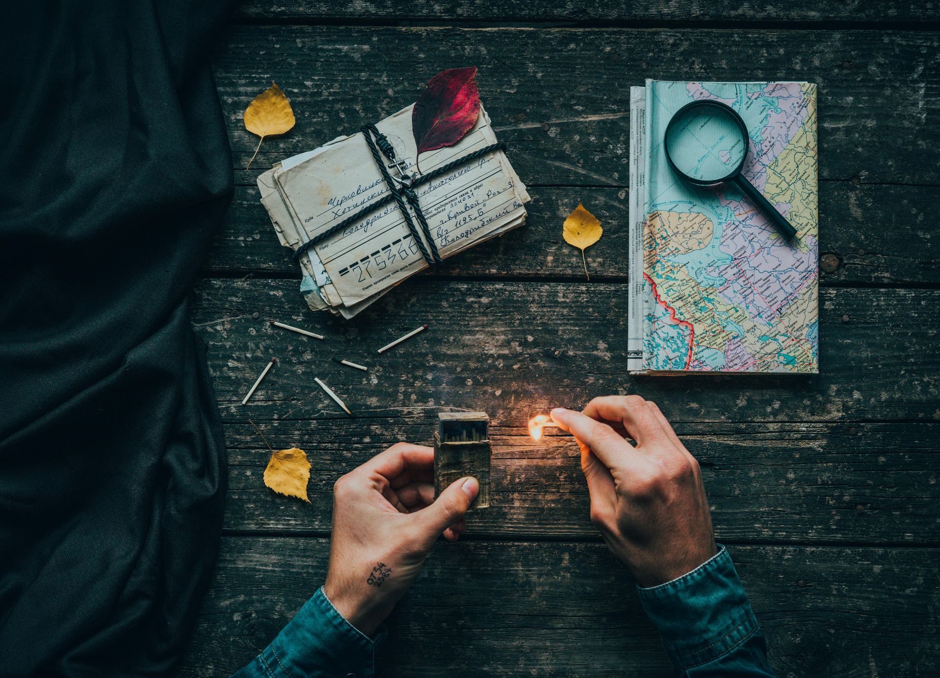 Hands lighting a match near a stack of papers tied with string, a map, and magnifying glass on a wooden surface.