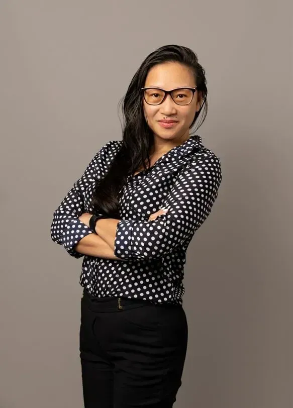 A woman wearing glasses and a polka dot shirt is standing with her arms crossed.