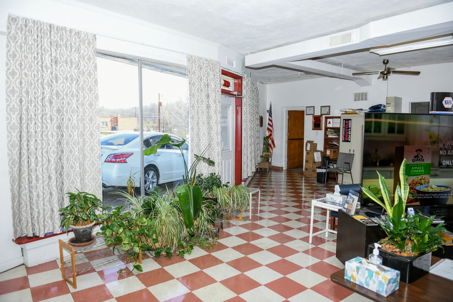 Bright plant-filled room with red-and-white checkered floor, large windows, and a parked car visible outside