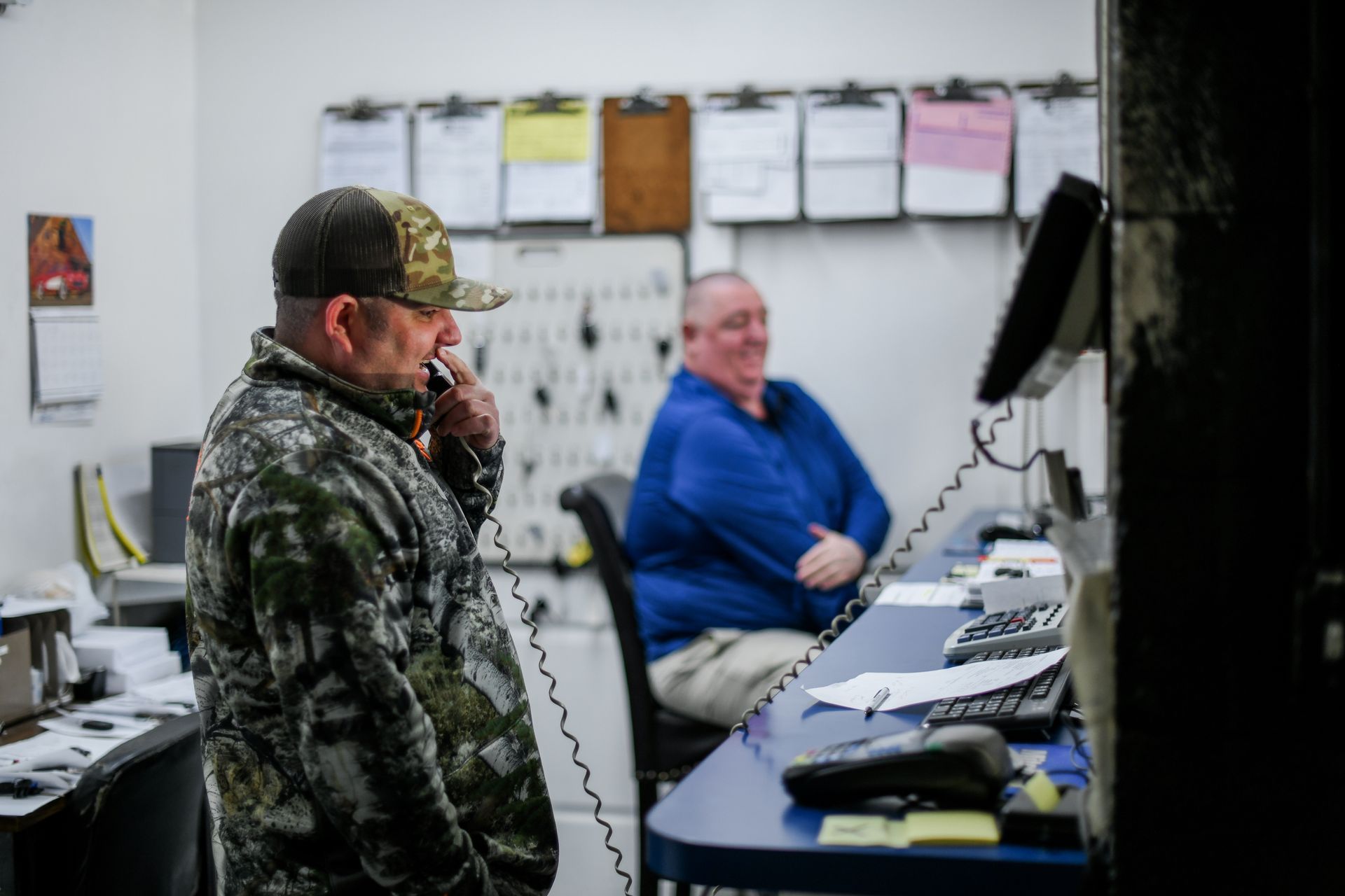 Two workers in a workshop, one standing in camouflage and one seated in blue at a desk with equipment.