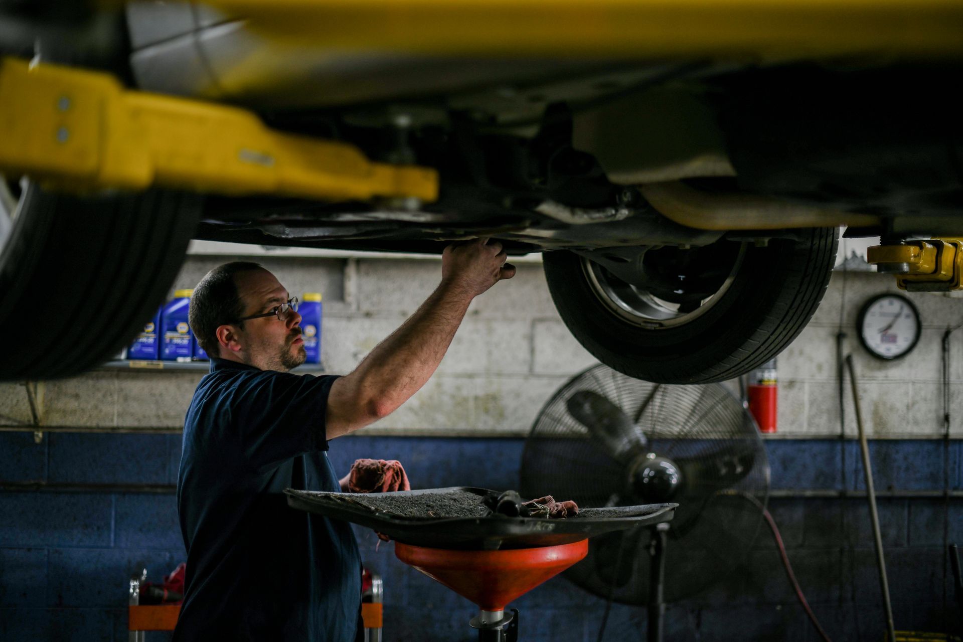 Mechanic inspecting a car’s undercarriage in a garage, standing by a red oil drain pan.