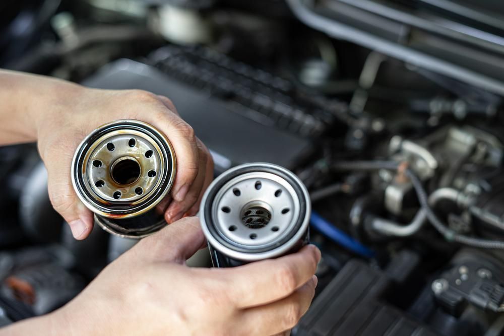 A Person is Holding Two Oil Filters in Their Hands in Front of a Car Engine — Ballina Bearing Supplies In Lismore, NSW
