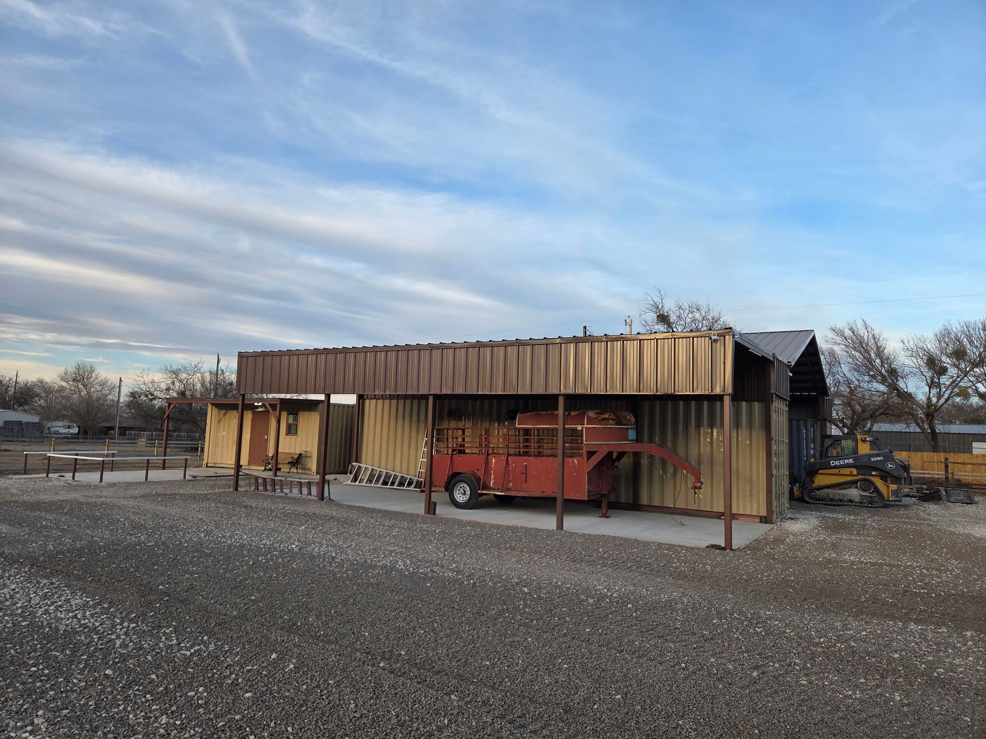 A red trailer parked under a brown-roofed shed on a gravel lot under a cloudy sky.