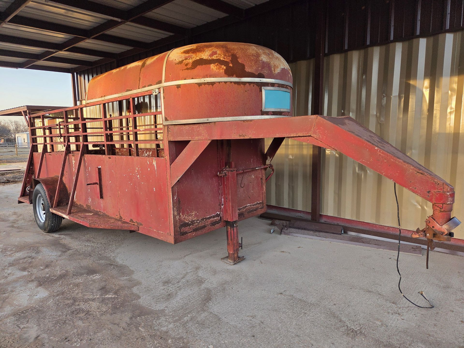 Red, rusty livestock trailer parked under a metal roof structure.