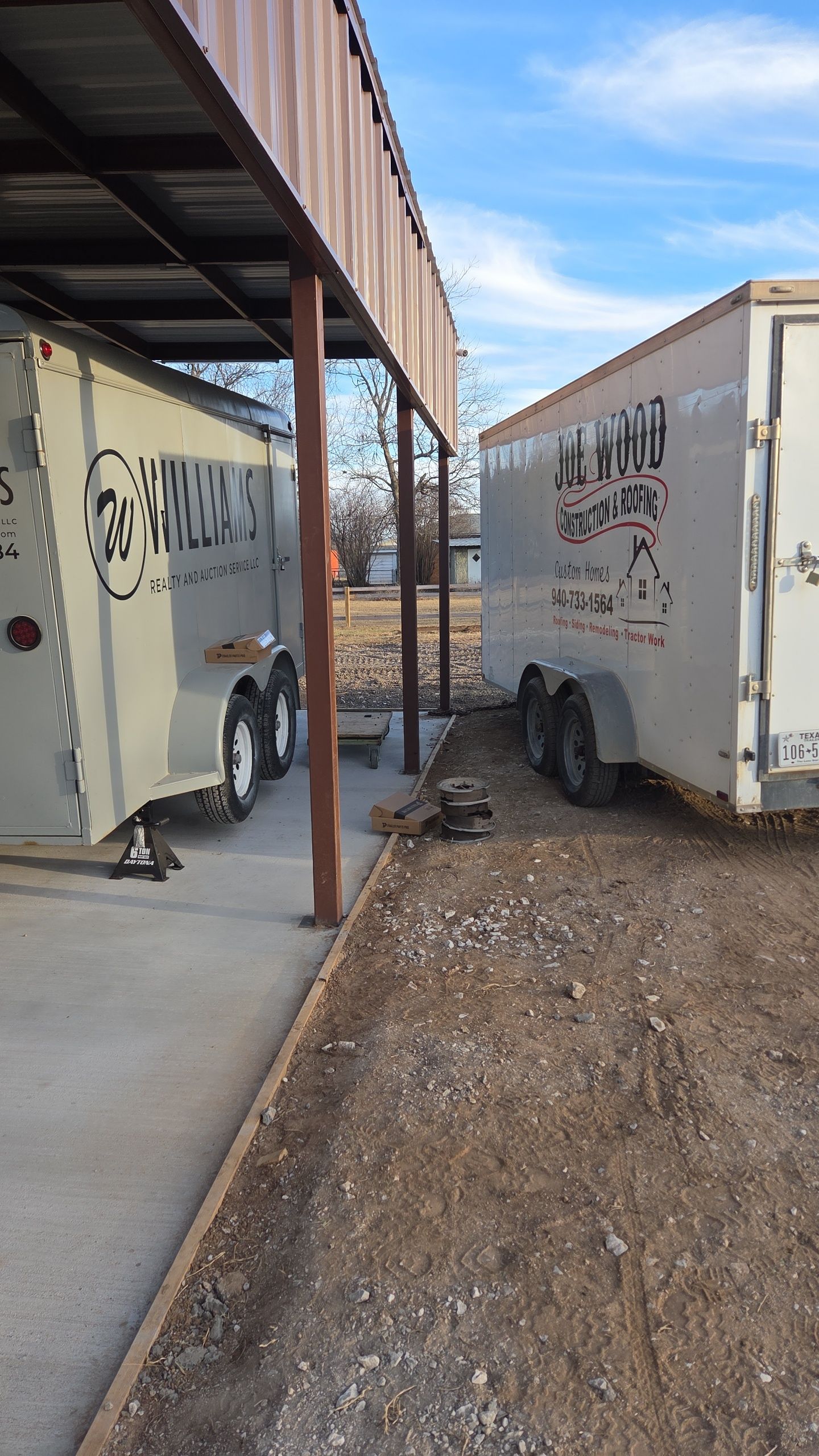 Two horse trailers parked under a brown awning, gravel ground, blue sky.
