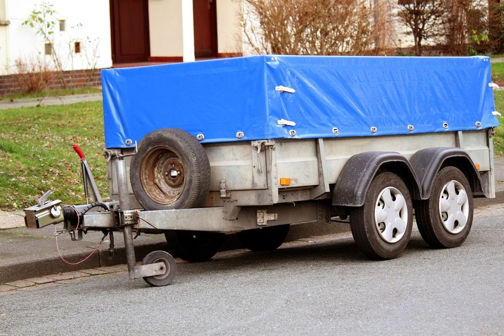 A blue-covered utility trailer parked on a street curb, with two tires and a spare.