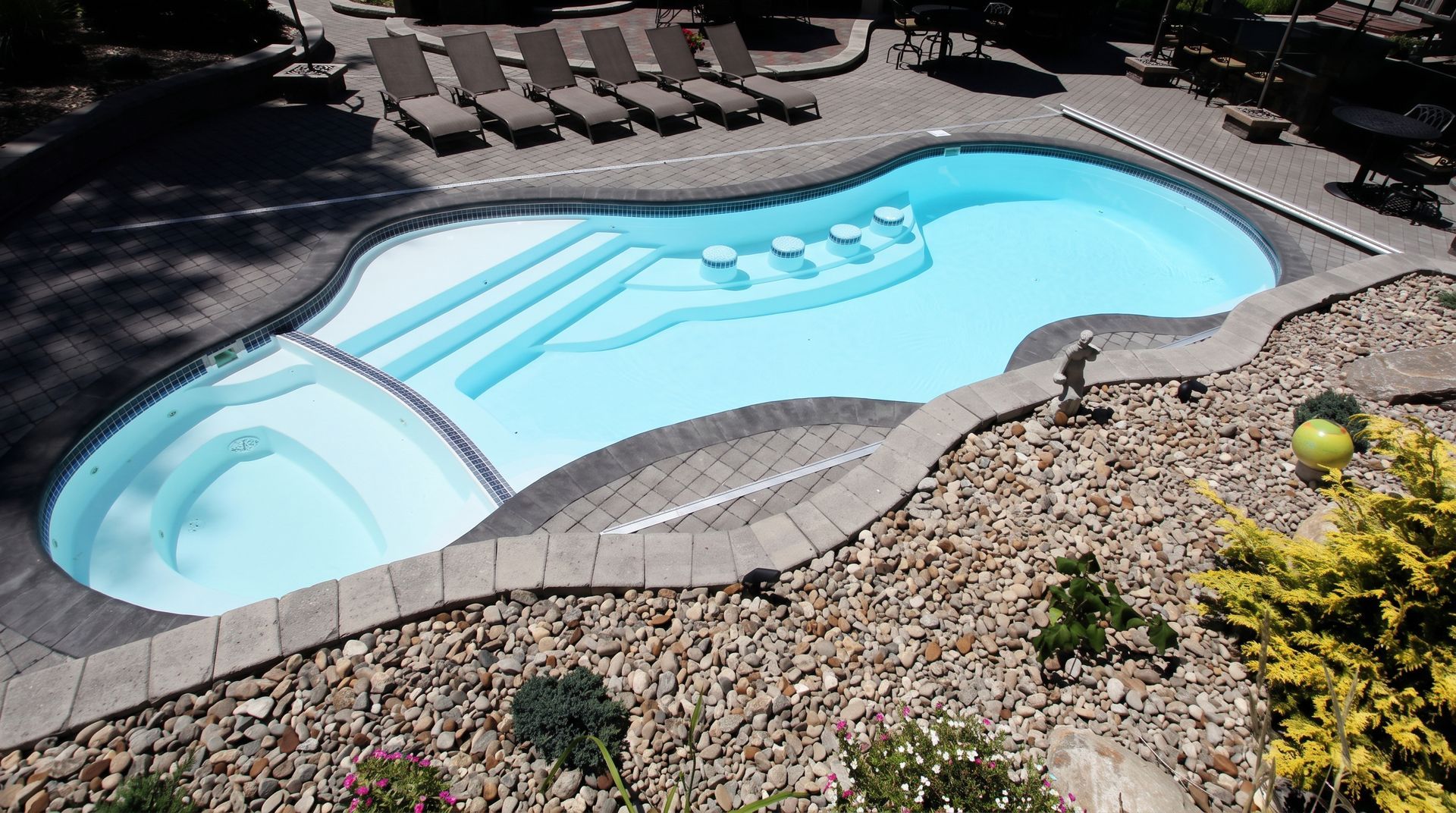 Kidney-shaped light blue swimming pool with built-in steps, surrounded by stone patio and gravel landscaping