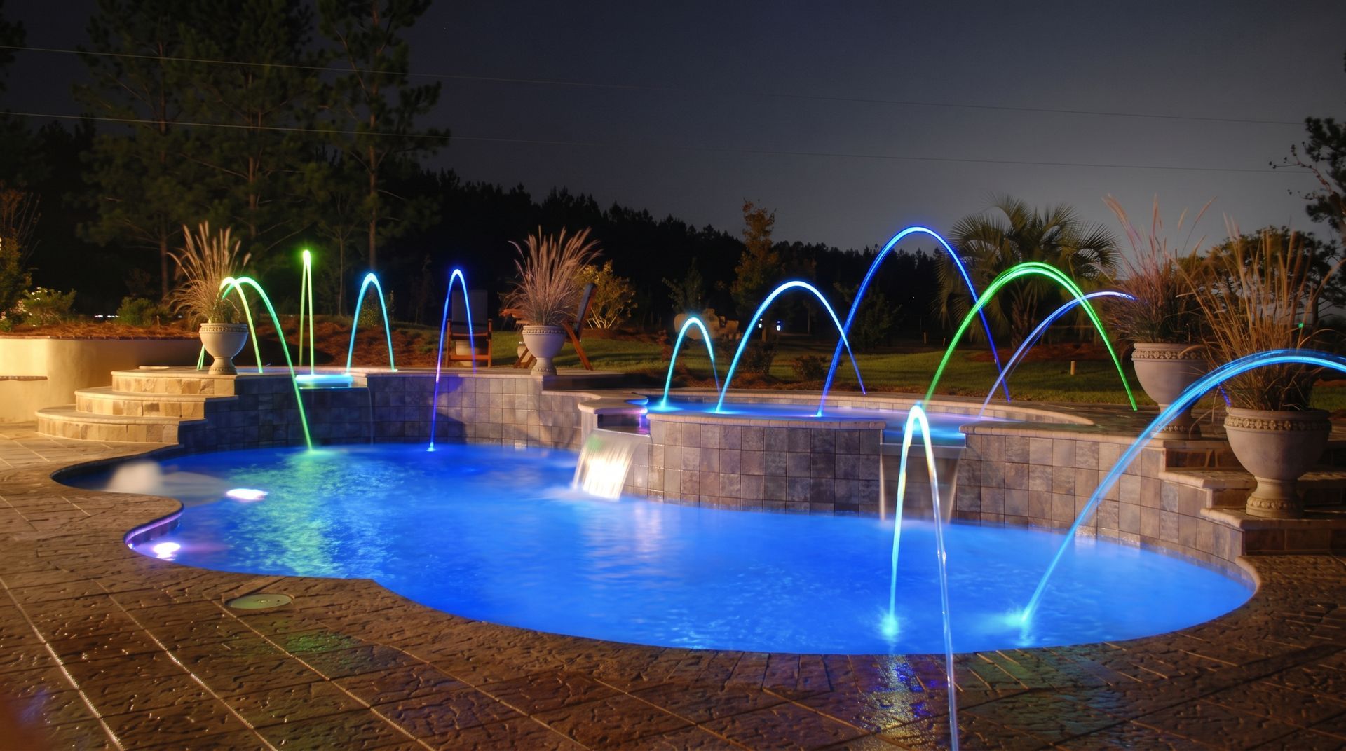 Illuminated backyard pool at night with colorful arcing fountains and blue water