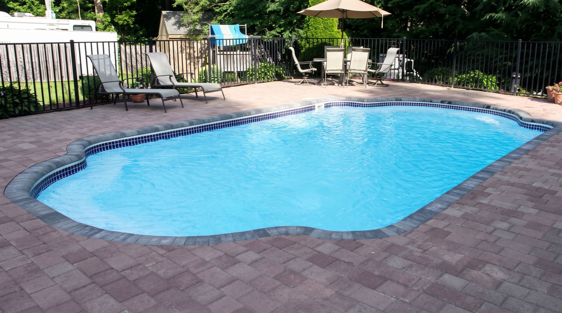 Backyard swimming pool with blue water, brick decking, and patio chairs in the background
