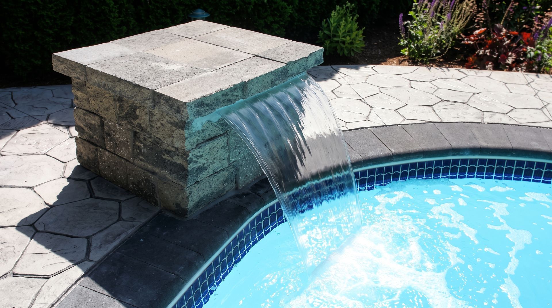 Stone waterfall feature pouring into a blue swimming pool beside a sunlit patio.