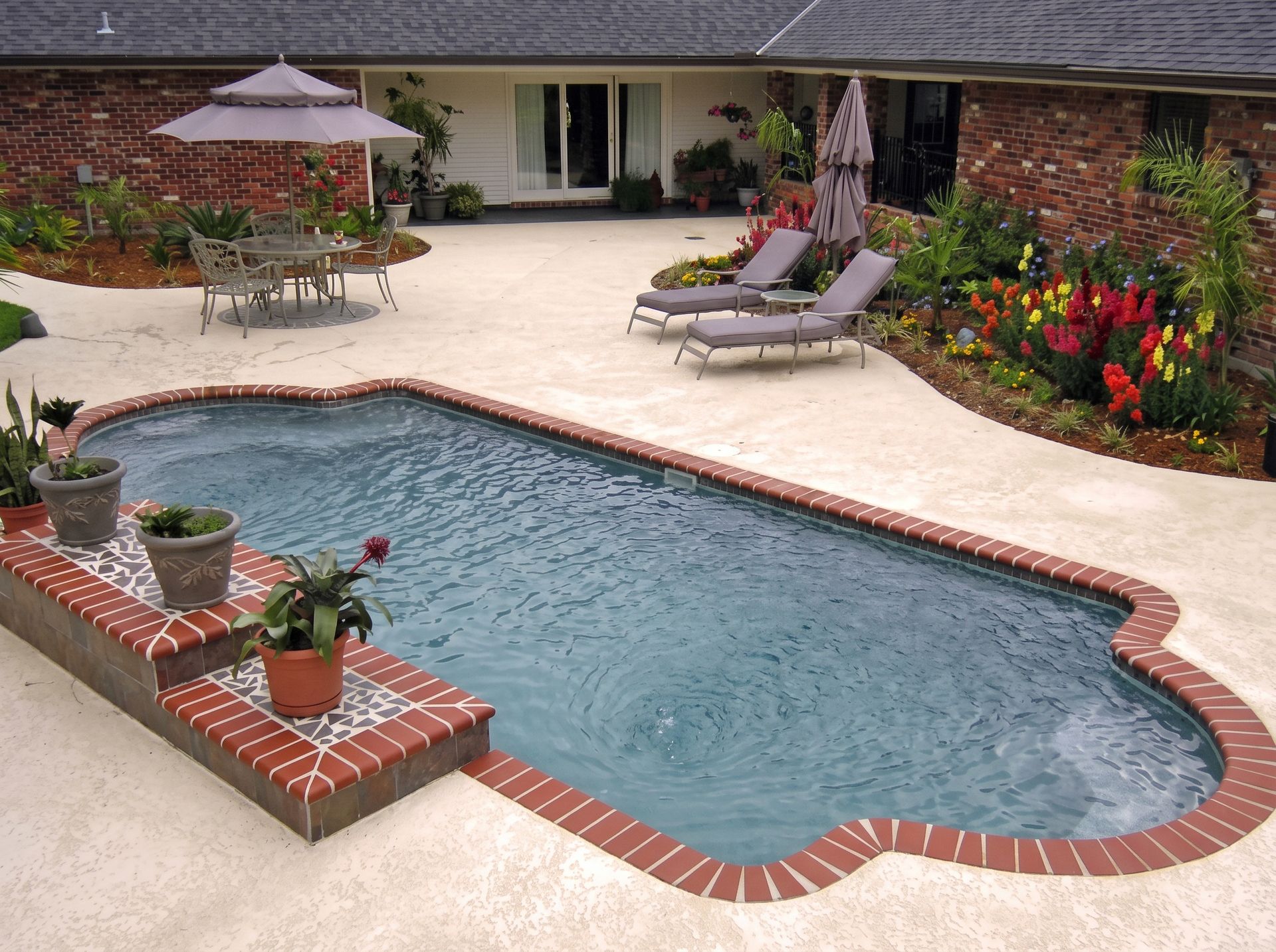 Backyard pool with brick trim, lounge chairs, umbrella, and flower beds beside a patio