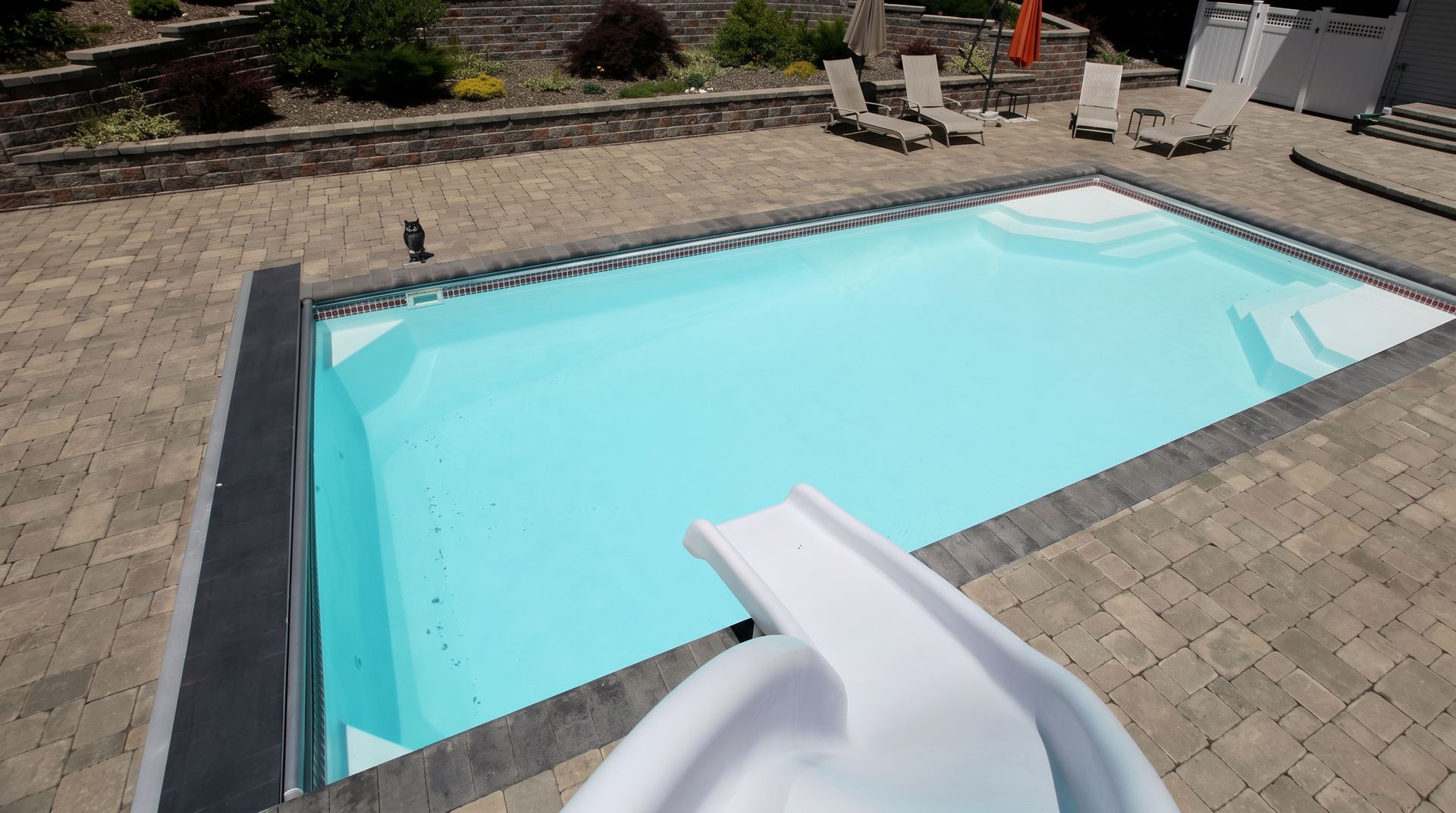 Backyard swimming pool with a white slide and stone paver deck in bright sunlight