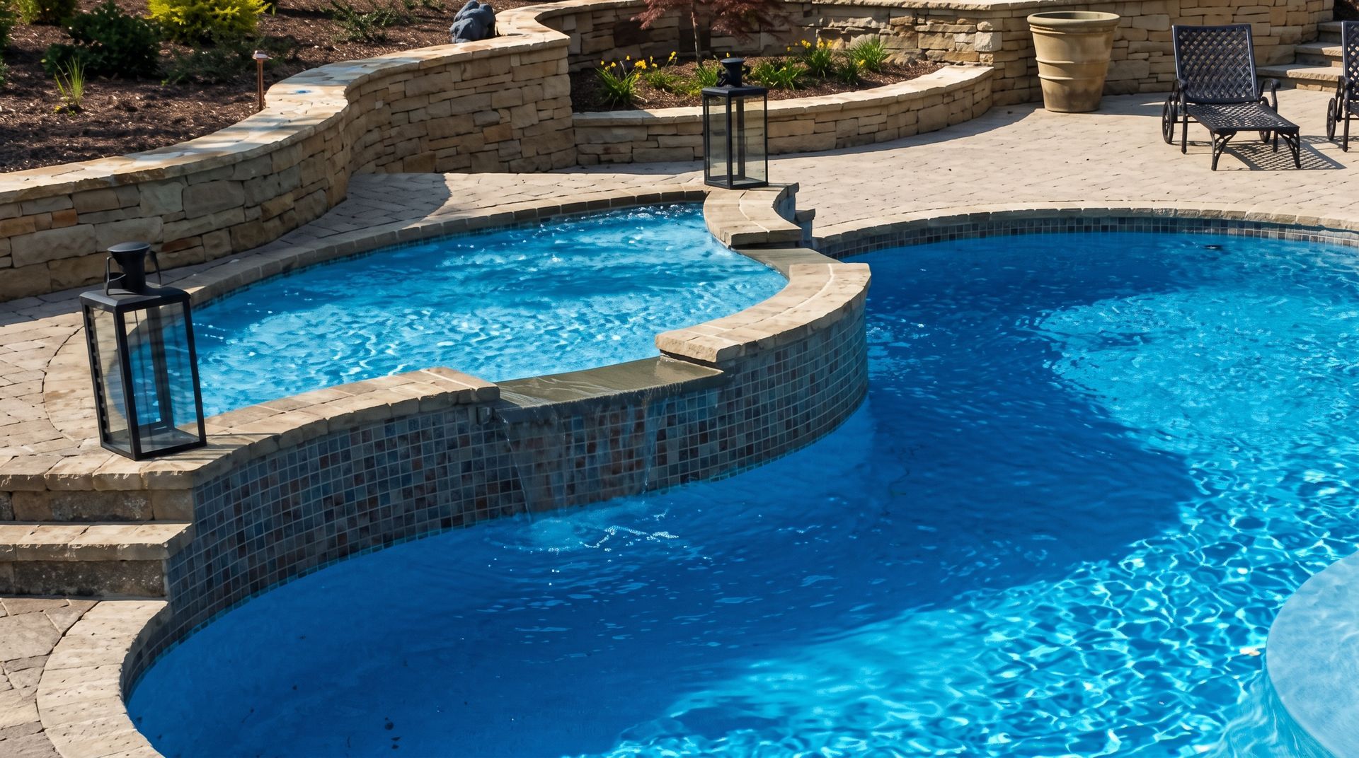 Curved backyard swimming pool with bright blue water, stone coping, and patio chairs in the background