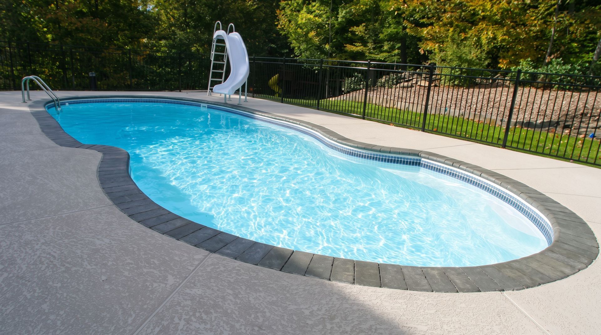 Kidney-shaped backyard swimming pool with a curved slide and dark gray concrete deck, bordered by a fence and trees