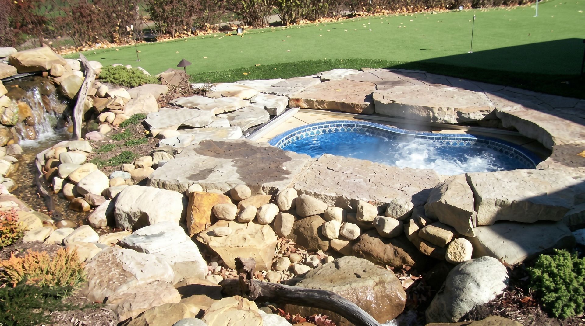Stone-lined backyard pond with a small waterfall and blue water, surrounded by rocks and lawn