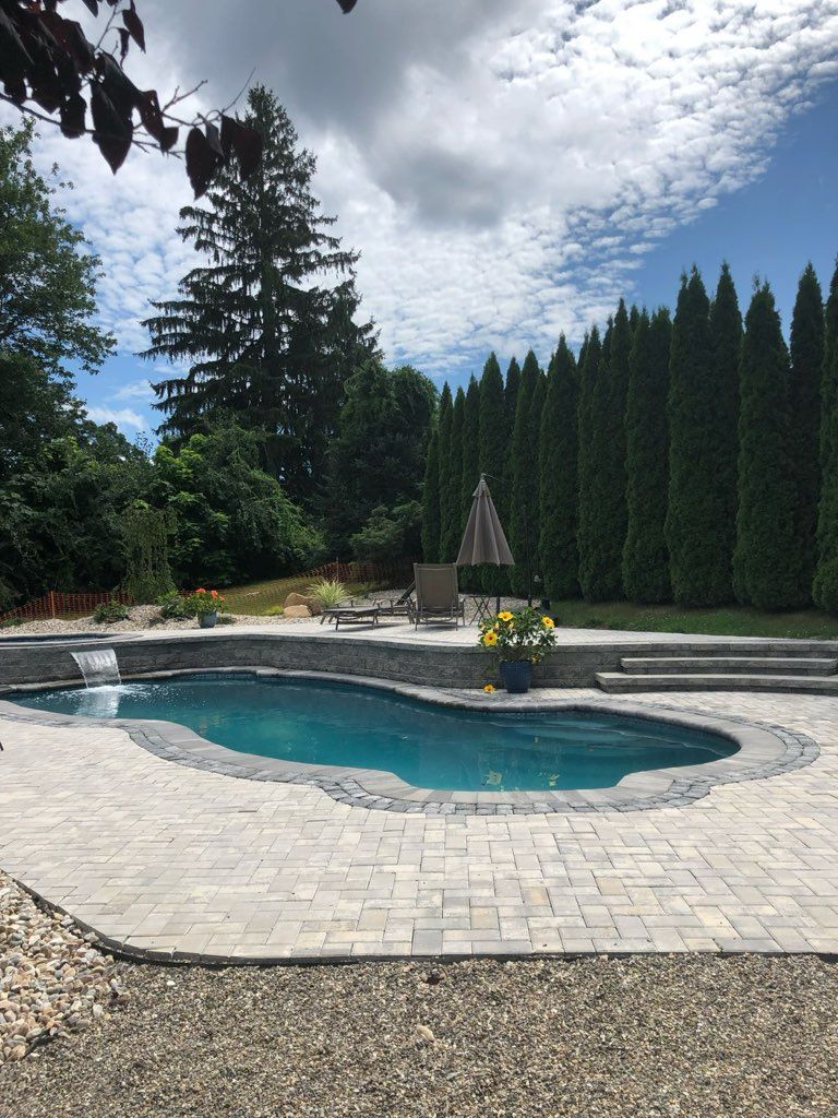 Backyard pool with stone deck, turquoise water, tall evergreens, and a central fountain under a cloudy sky