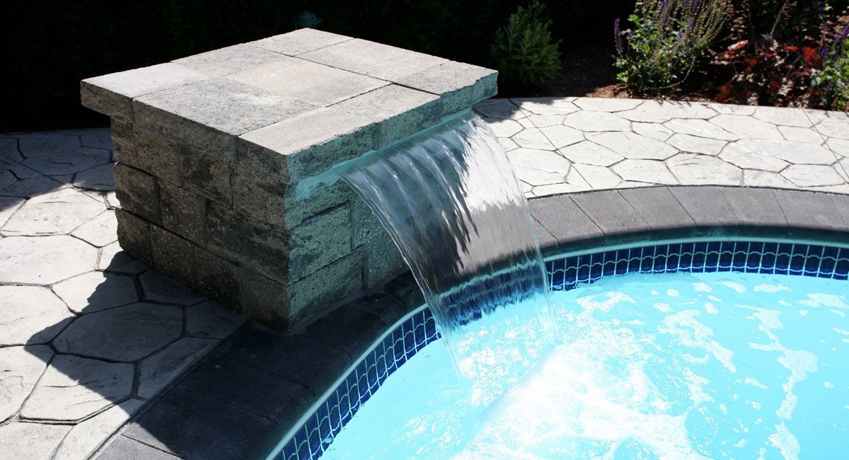 Stone waterfall spilling into a blue swimming pool beside a sunlit patio