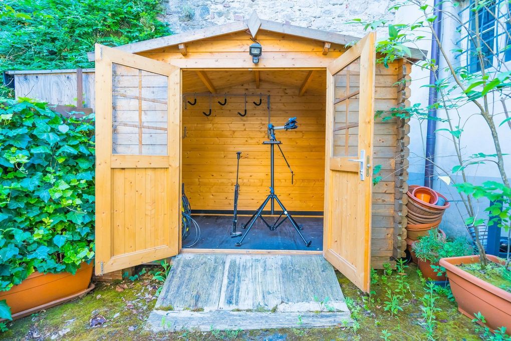 Casetta da giardino in legno aperta con doppie porte, vuota all'interno ad eccezione di un treppiede sul pavimento.