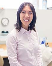 A woman in a pink shirt is standing in front of a desk and smiling.