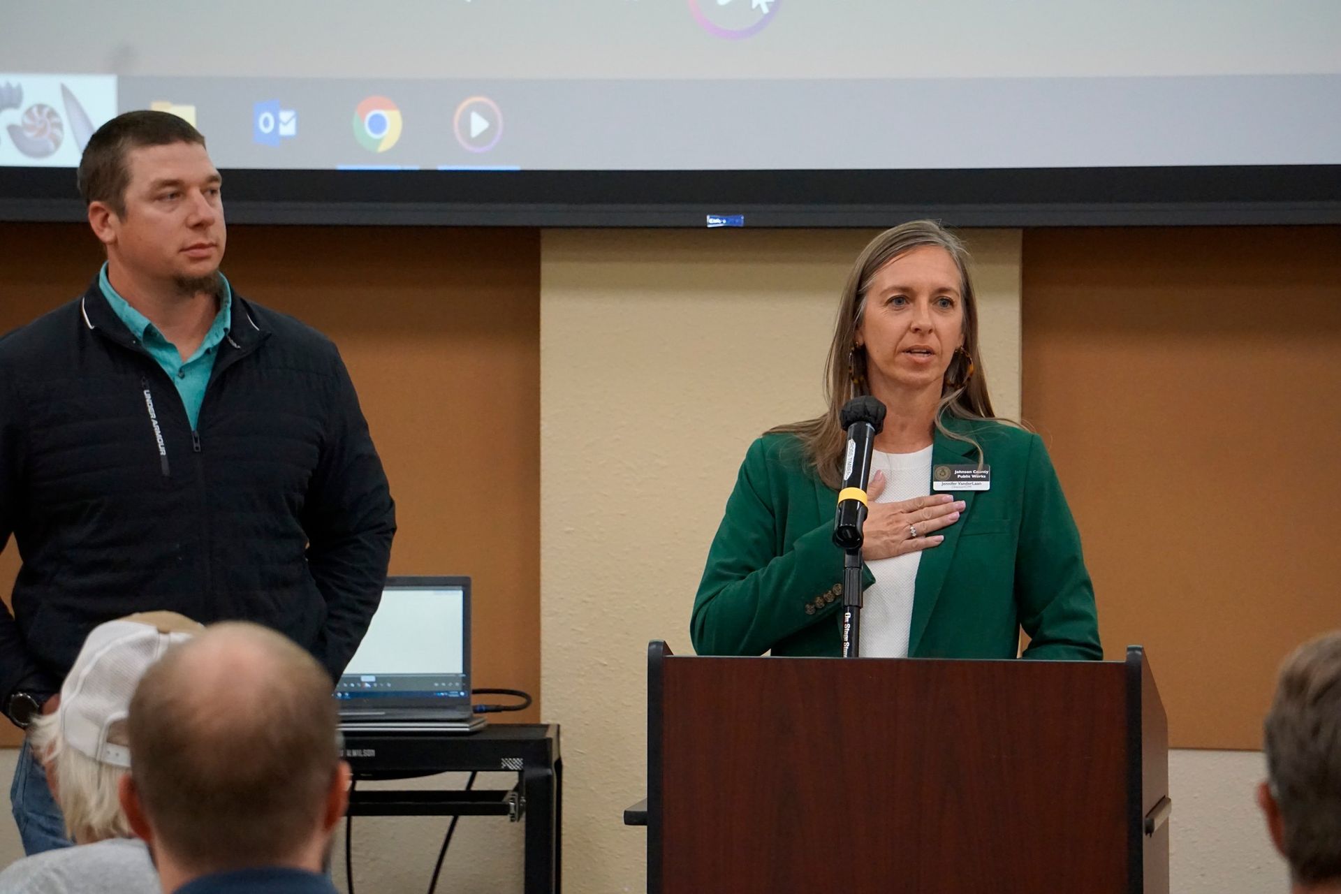 Two presenters stand at a podium during a presentation, with one speaking and a projected screen behind them.