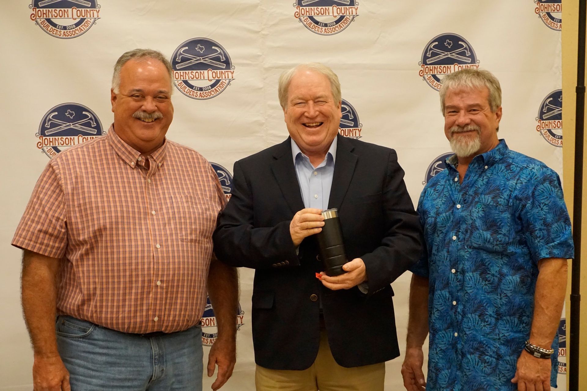 Three men pose at an event backdrop, smiling, with the center man holding a trophy.
