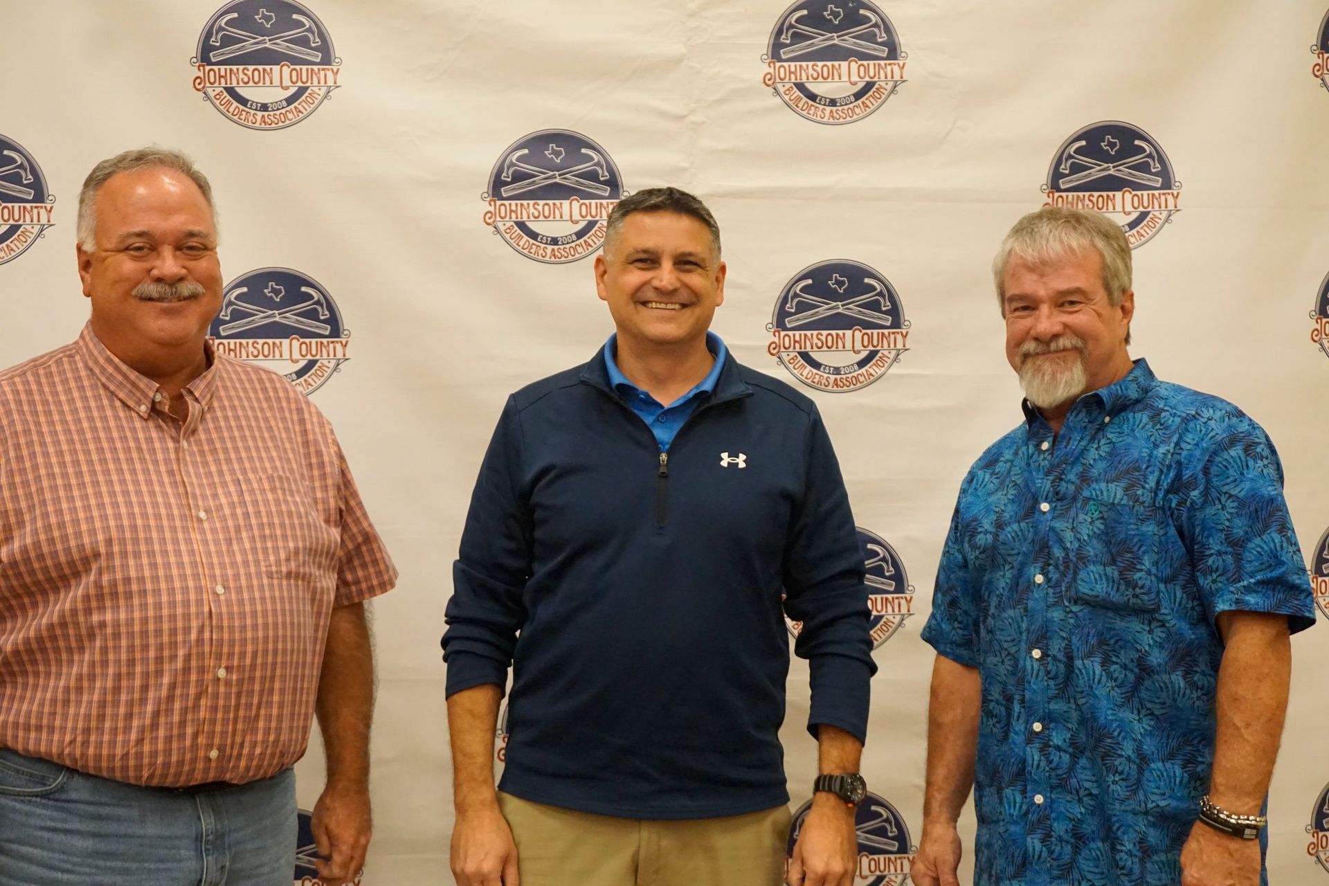 Three men standing in front of a logo backdrop, smiling at an indoor event.