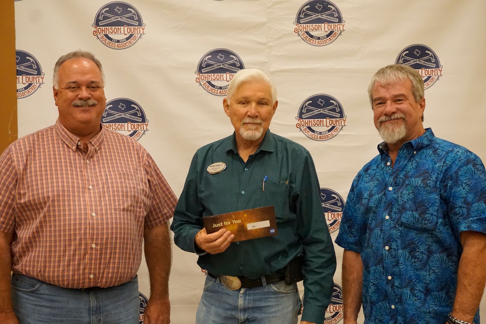 Three men pose in front of a logo backdrop, with the center man holding a plaque.