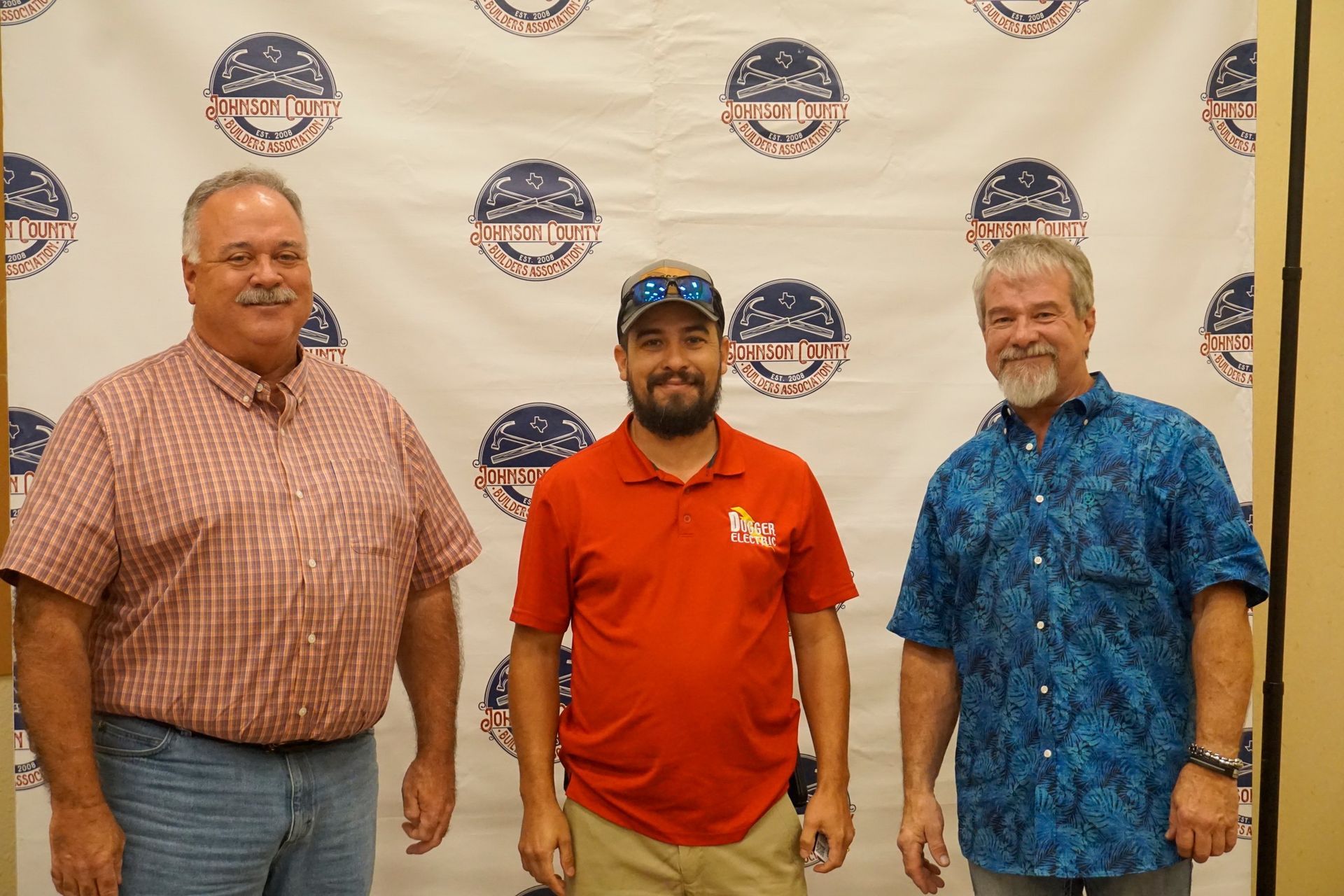 Three men stand posing in front of a logo backdrop, wearing red, plaid, and blue shirts.