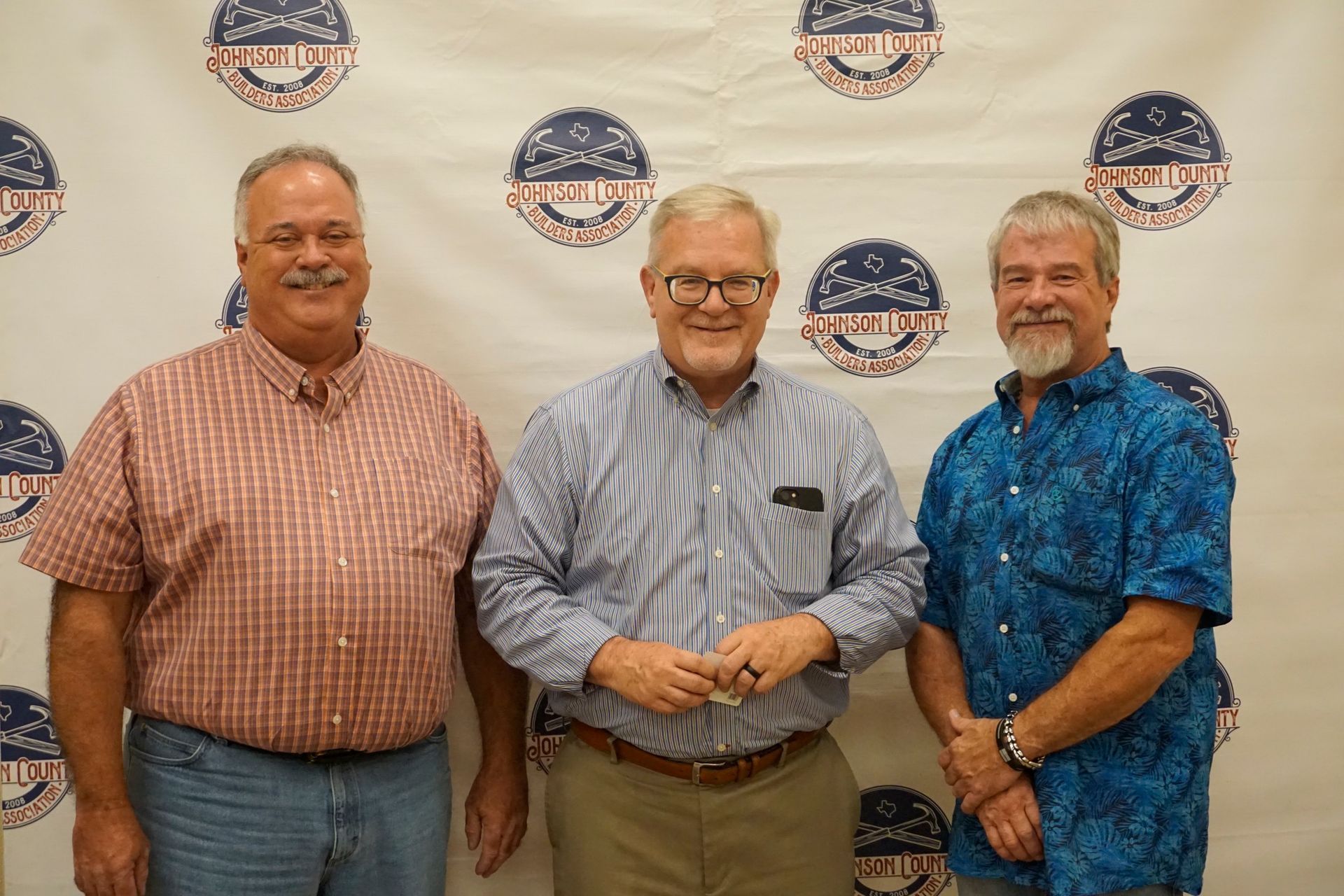Three men standing together and smiling at a branded backdrop, wearing casual button-up shirts.