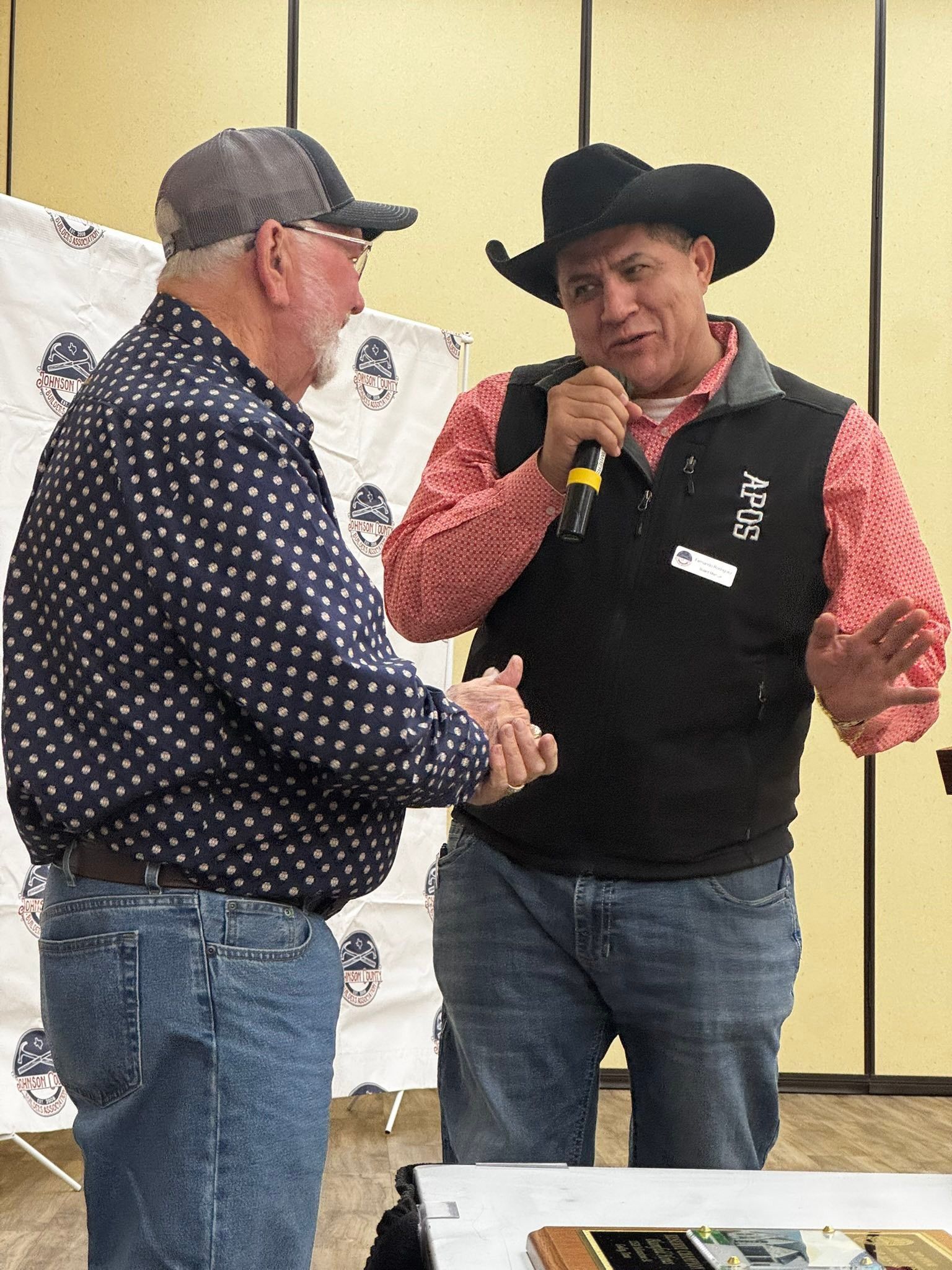 Two men shaking hands during a conversation, one holding a microphone, indoors.