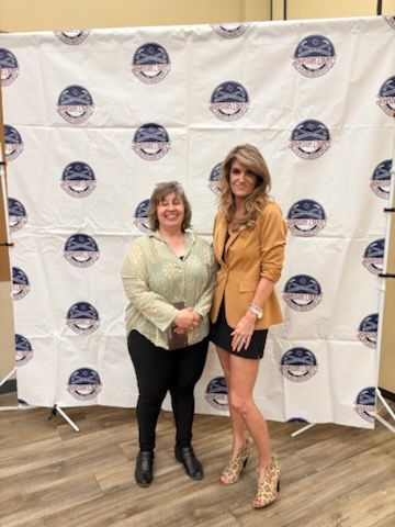 Two women posing in front of a step-and-repeat banner, one in a blazer and one in an orange top and black skirt.