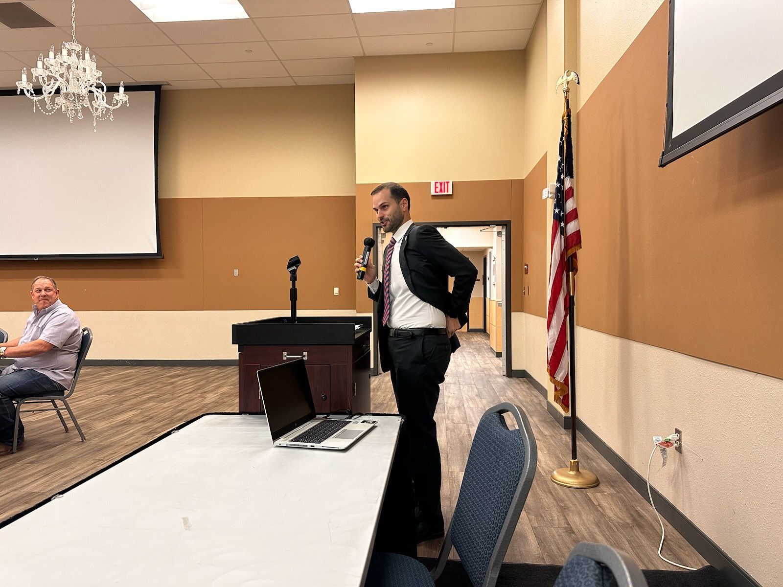 A man in a suit is standing in front of a microphone in a conference room.