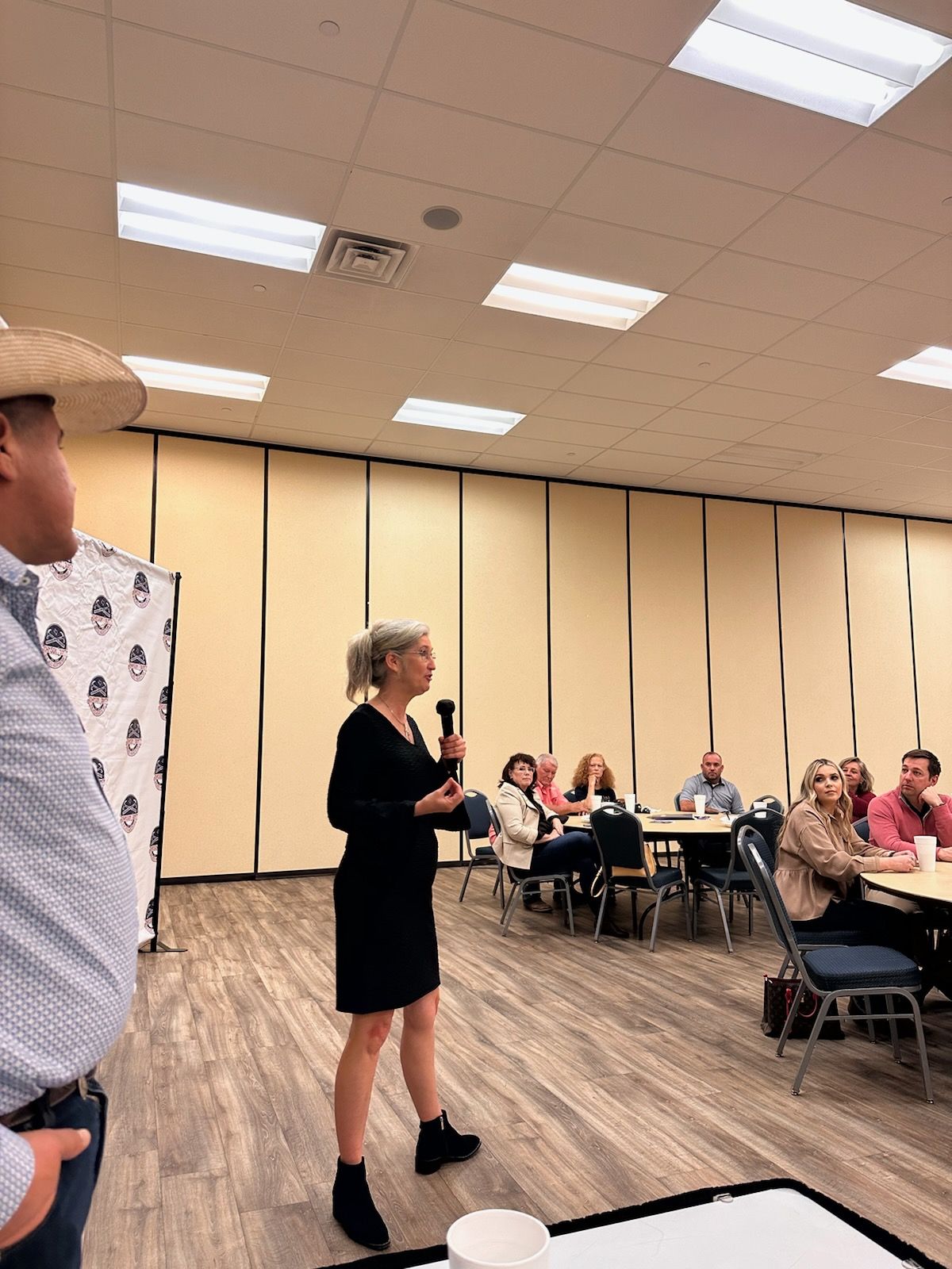 A woman is standing in front of a microphone in a room with people sitting at tables.