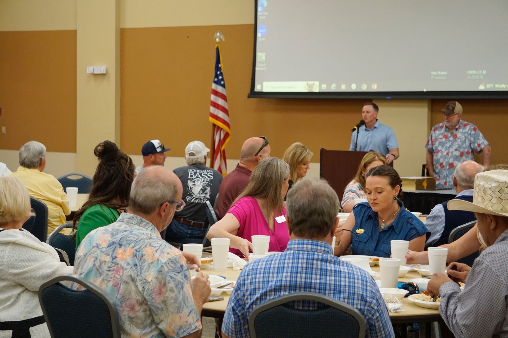 A group of people are sitting at tables in front of a projector screen.
