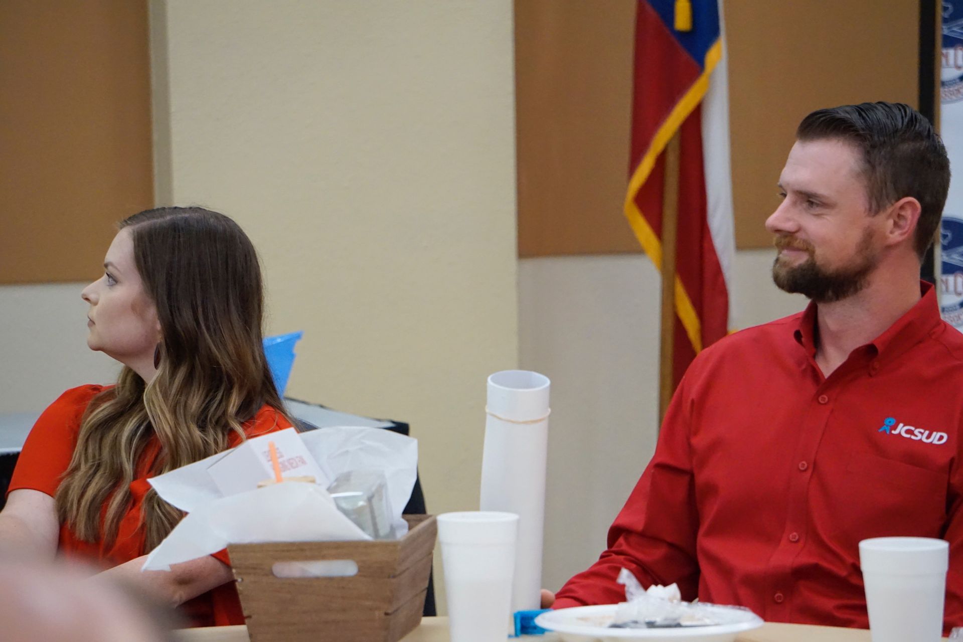 A man and a woman are sitting at a table in front of a flag.