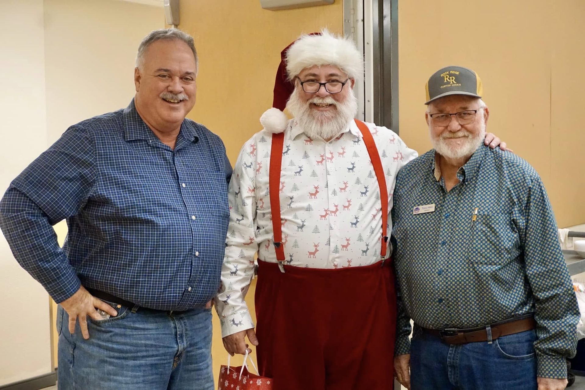 Three men posing indoors, one dressed as Santa in red suspenders and hat, smiling at the camera.
