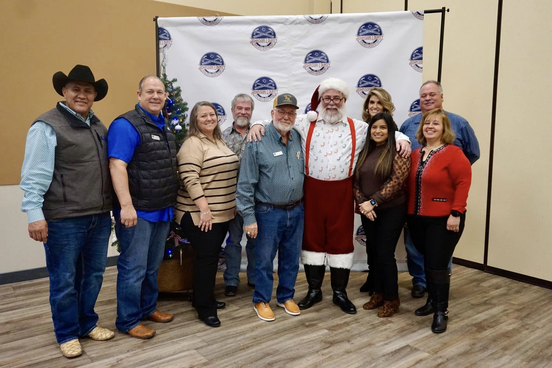 Group posing indoors at a holiday event, with one person dressed as Santa in front of a branded backdrop.