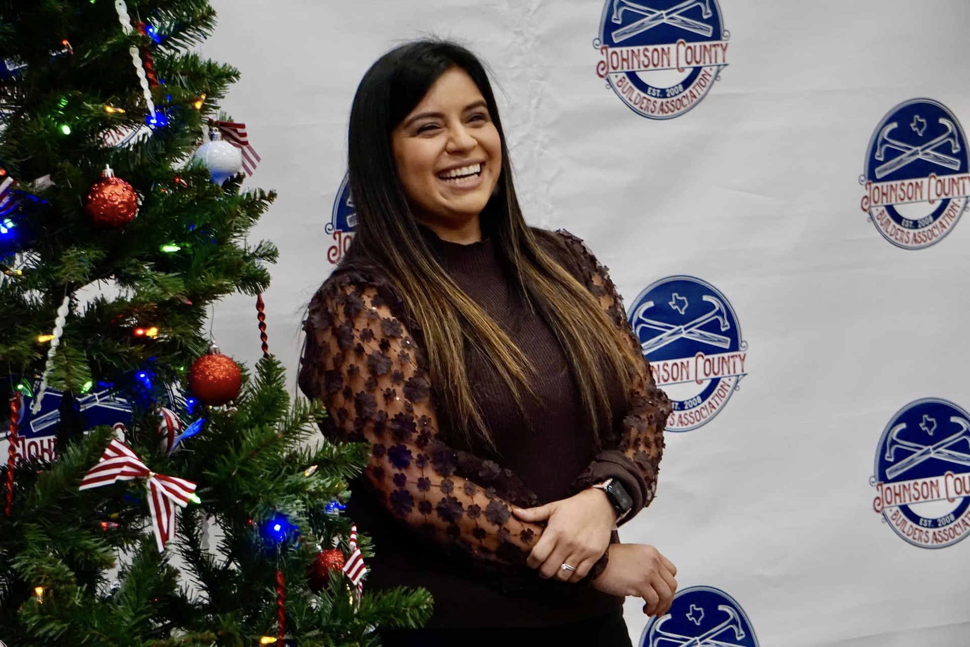 Smiling woman in a patterned dress stands beside a decorated Christmas tree at a holiday event backdrop