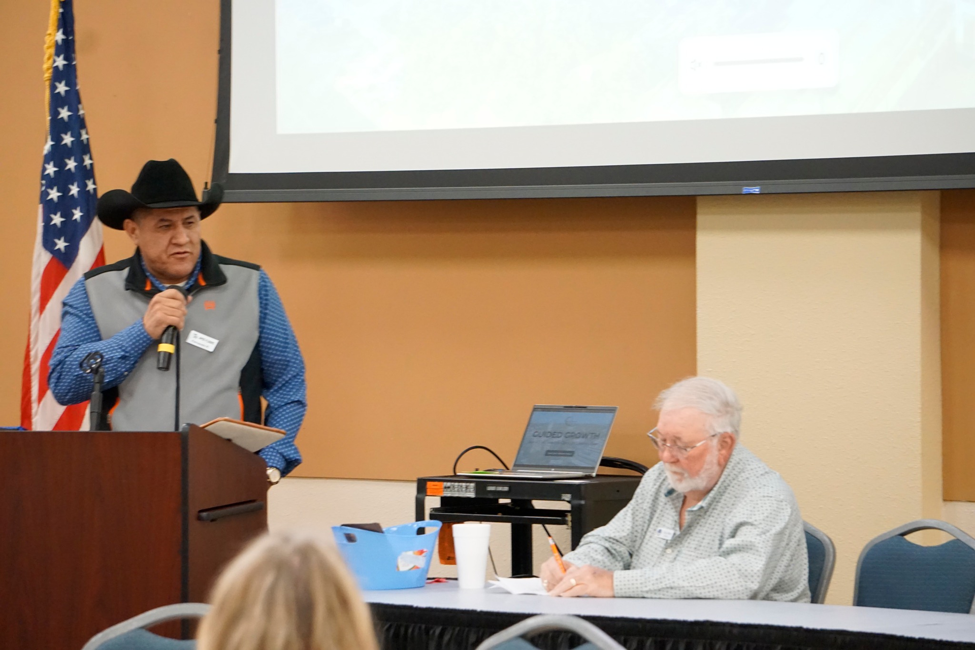 Speaker at podium addresses audience while seated man works at table in meeting room