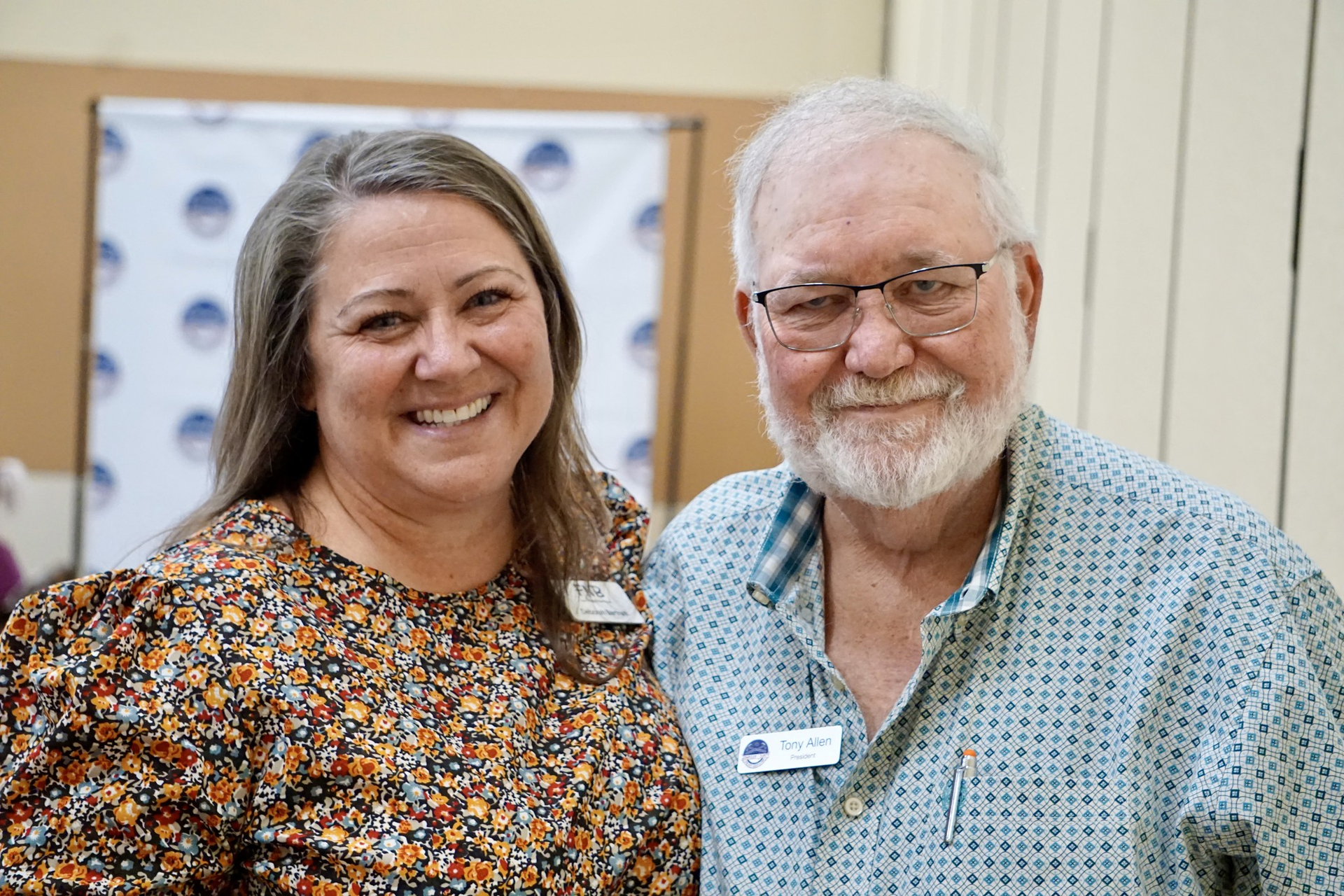 Two smiling people standing indoors, wearing name tags and patterned shirts.