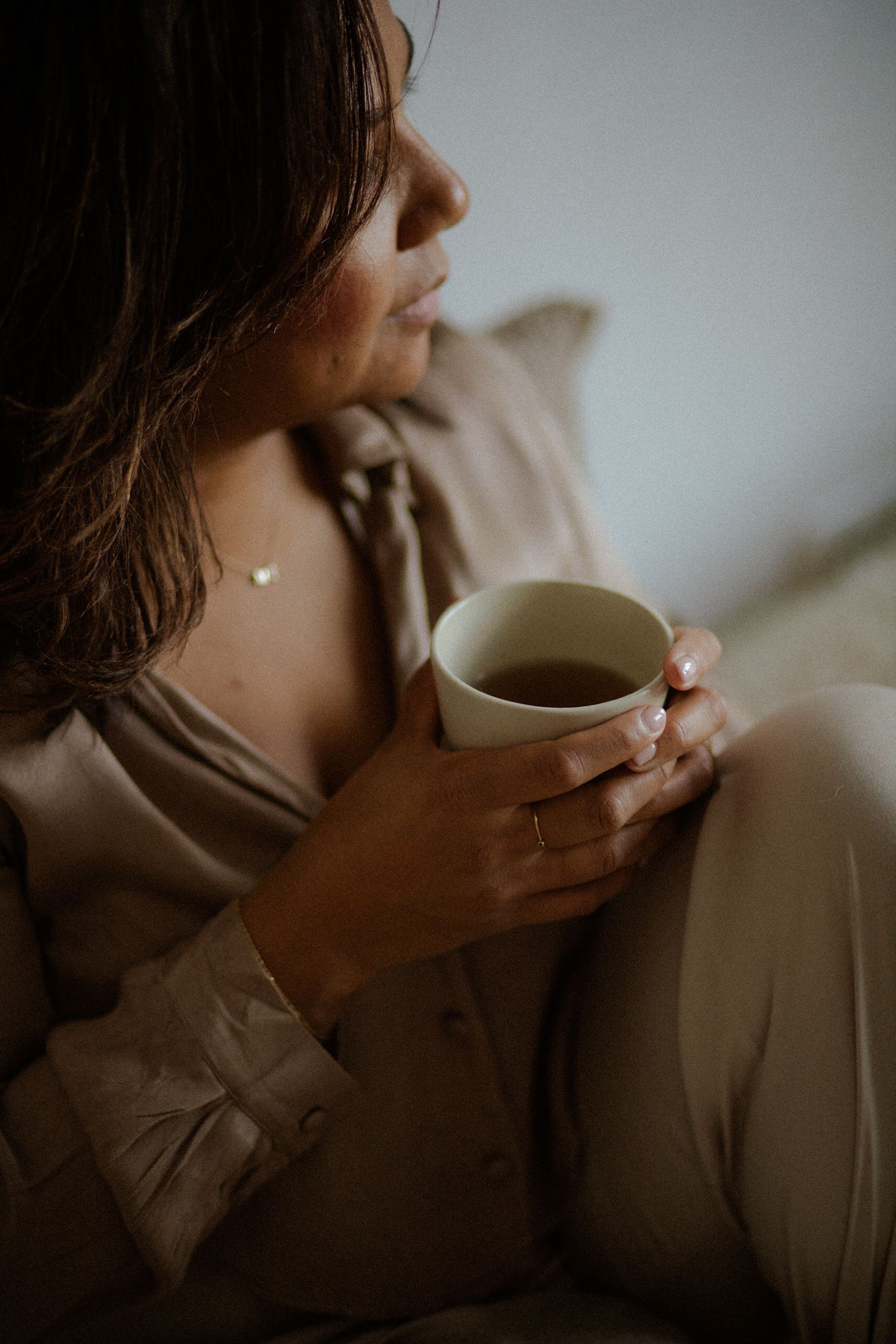 Woman holding a mug, gazing off. Wearing tan button-up shirt and pants, simple gold necklace. Light, indoor setting.