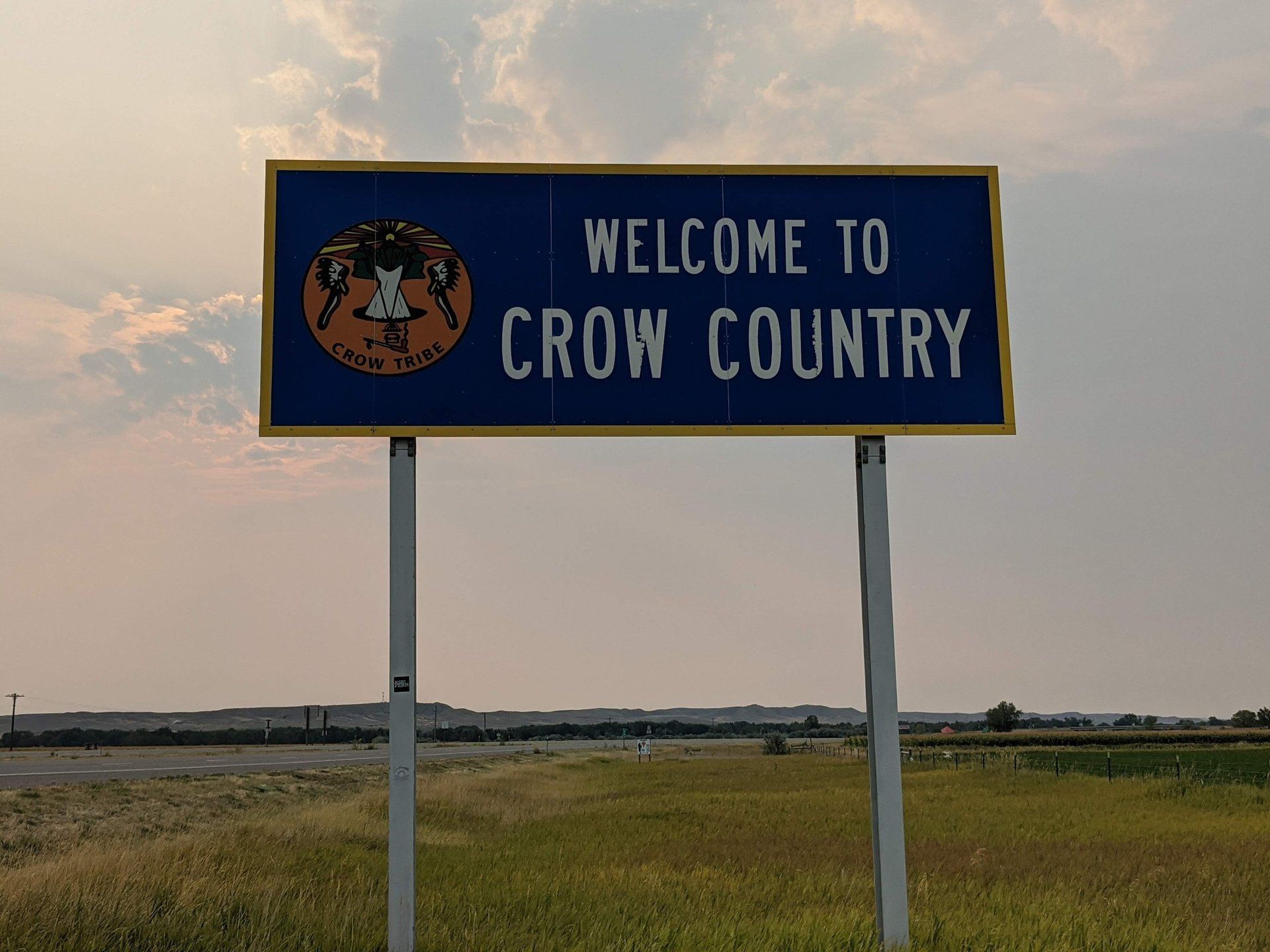 Blue roadside sign reading “Welcome to Crow Country” in a grassy field at sunset