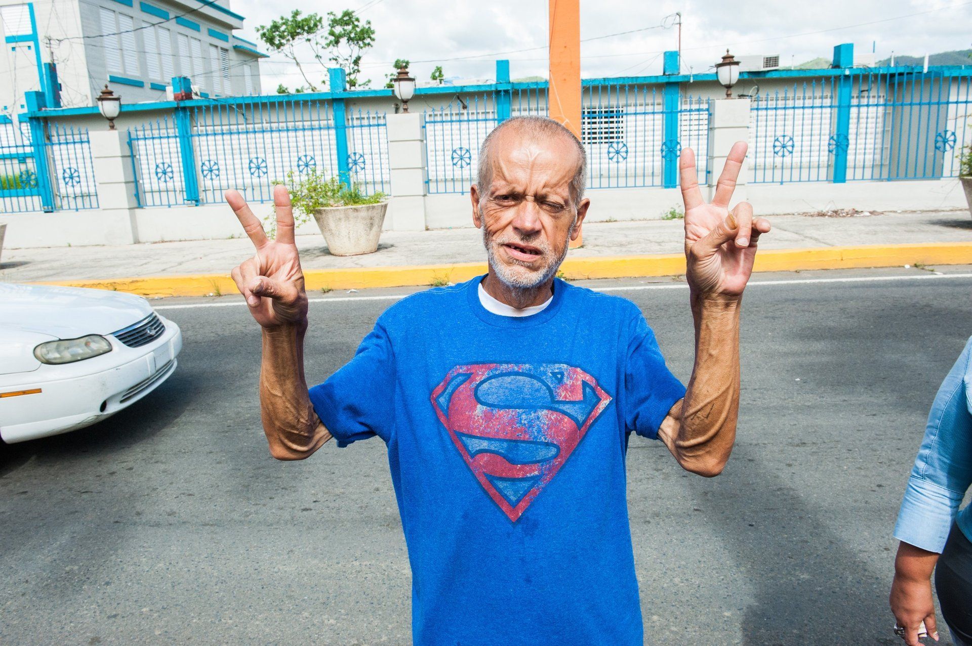 Man in blue Superman T-shirt holding up peace signs on a street with cars and a blue fence behind him